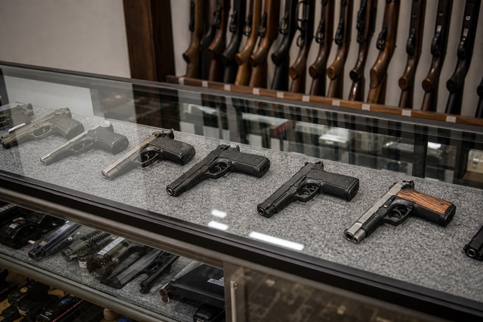 AI-generated illustration of three-quarter close view of a clean gun-store glass display counter containing neatly spaced unloaded handguns on gray felt, secured long-gun racks behind, realistic reflections
