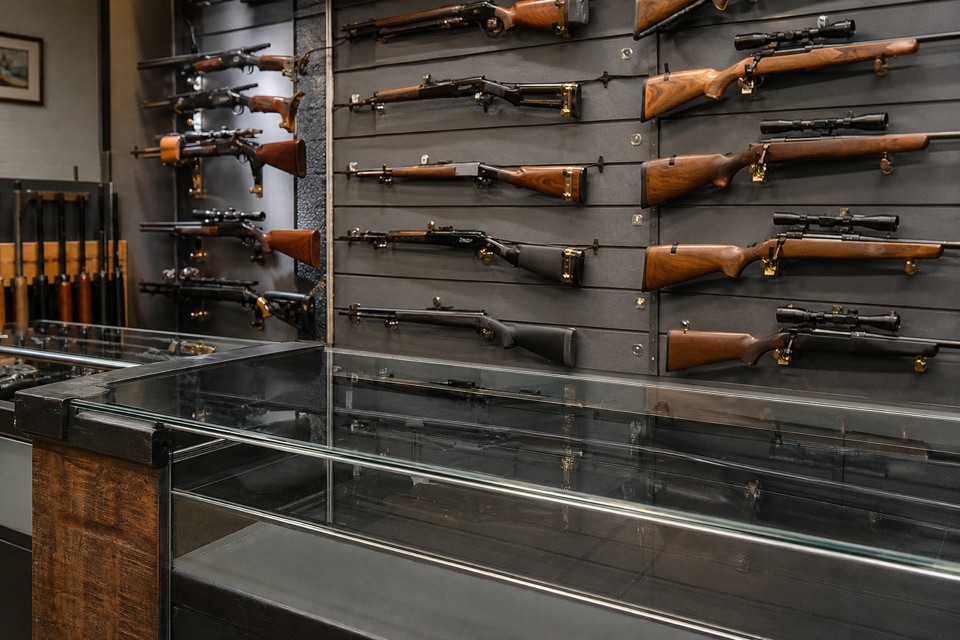 AI-generated illustration of medium-wide gun-shop counter scene, empty clean glass counter foreground, clearly visible secured civilian sporting rifles on slate-gray wall behind, subtle brass highlights