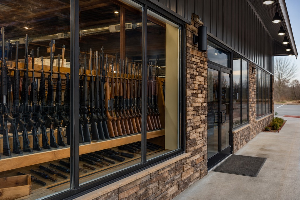 AI-generated illustration of exterior architectural photograph of a suburban American firearm dealer in a clean low-rise retail building, secure windows showing rifle racks, neutral facade, no readable branding