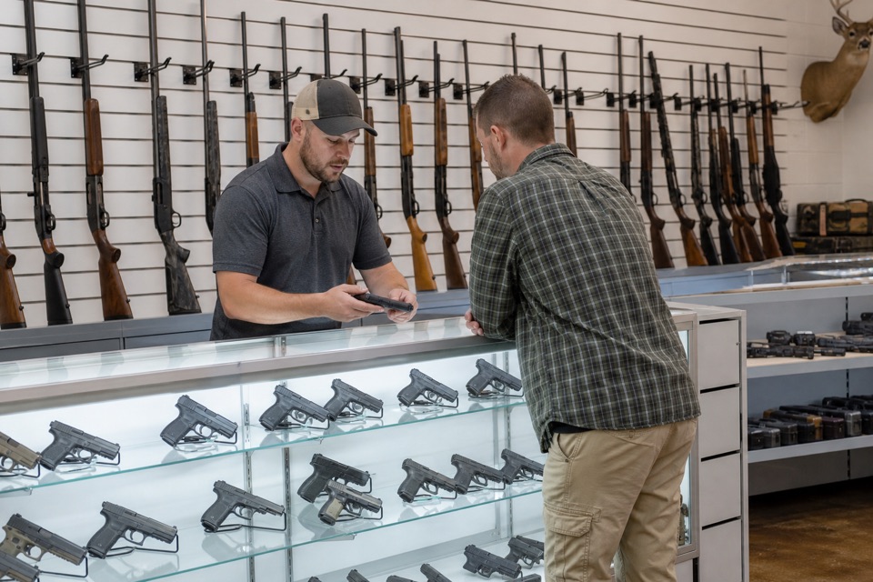 AI-generated illustration of a bright licensed firearm shop with white slatwall, orderly rows of secured sporting rifles and a central locked handgun display island, crisp natural photographic color