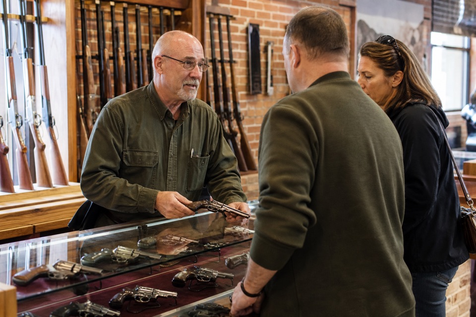 AI-generated illustration of established family-run firearms shop with red-brick interior, oak long-gun wall racks and glass revolver display counters, warm natural light, no logos