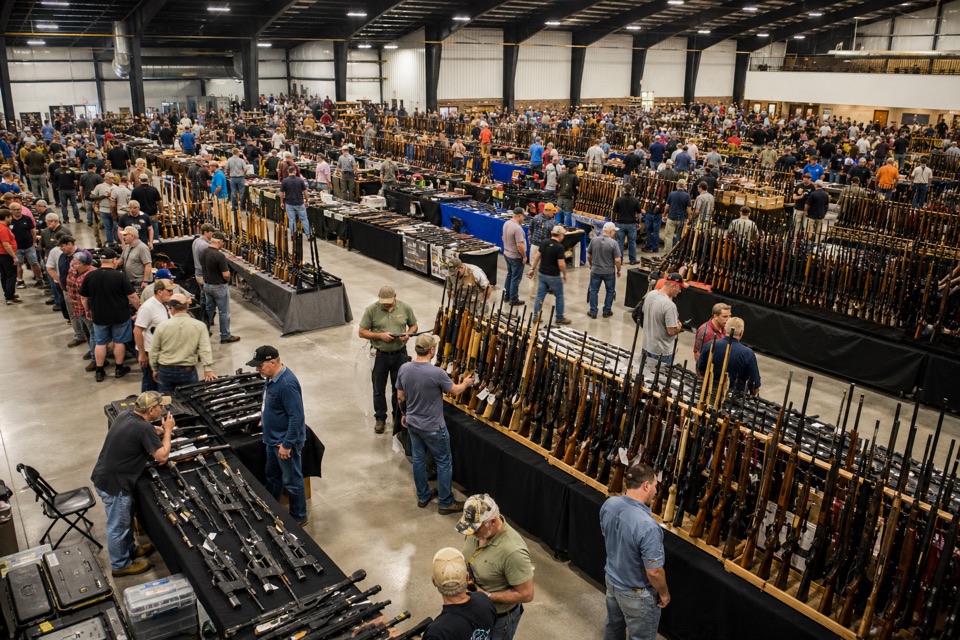 AI-generated illustration of slightly elevated wide overview of a large American gun show, unmistakable rows of firearm display tables, bright modern steel-roof exhibition building, adults browsing