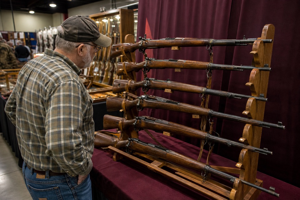 AI-generated illustration of a collectors booth at a gun show with vintage wood-stock bolt-action rifles secured on display stands, warm amber hall light and maroon backdrop