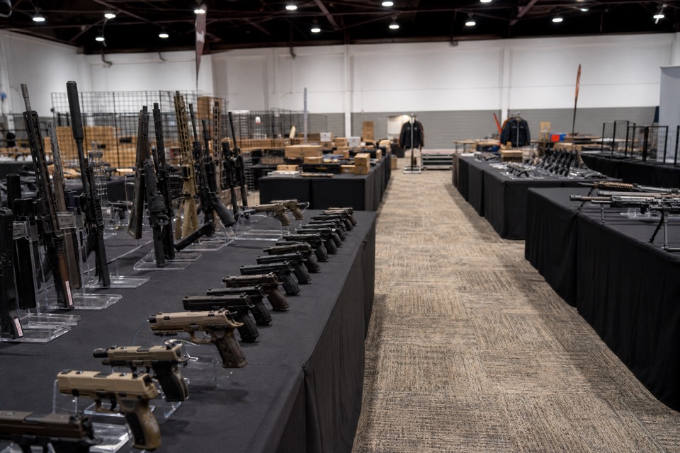 AI-generated illustration of early setup at a gun show before opening, orderly black-covered vendor tables with secured unloaded firearm displays and empty carpeted aisles, crisp architectural view