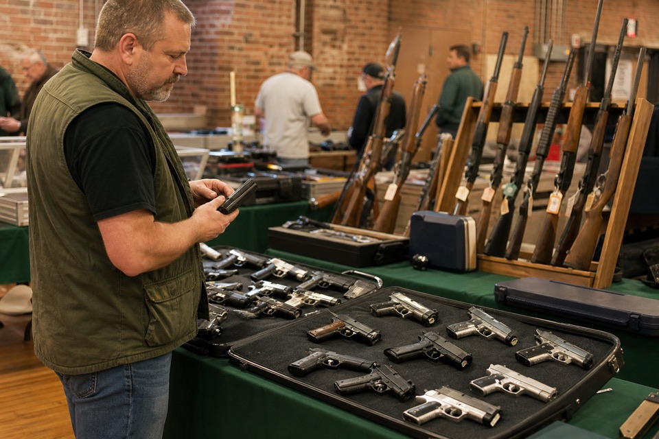 AI-generated illustration of medium shot of a local gun-show corner table with unloaded sporting handguns and rifles in padded displays, forest-green cloth, adult vendor, softly blurred red-brick hall